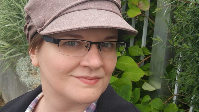 Photo shows head and shoulders of white person with blue eyes and dark grey glasses, wearing a dark coat, pastel plaid shirt, and a beige hat with a brim and a button on the side. Alix stands in front of a wall out of which plants are growing.
