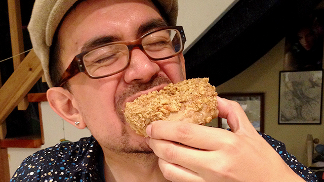 Photo shows a half-Filipino person with a moustache wearing a spotted blue shirt, a beige hat and tortoise-shell brown glasses. Chris is biting into a texture doughnut.