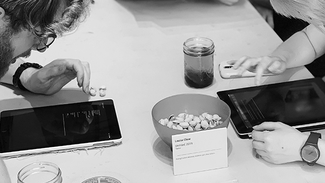 Photo description: A black and white image showing 2 people leaning over tablets, across from each other at a table. Between the tablets is a bowl of pistachio nuts, a glass of liquid, and a white title card for the project.