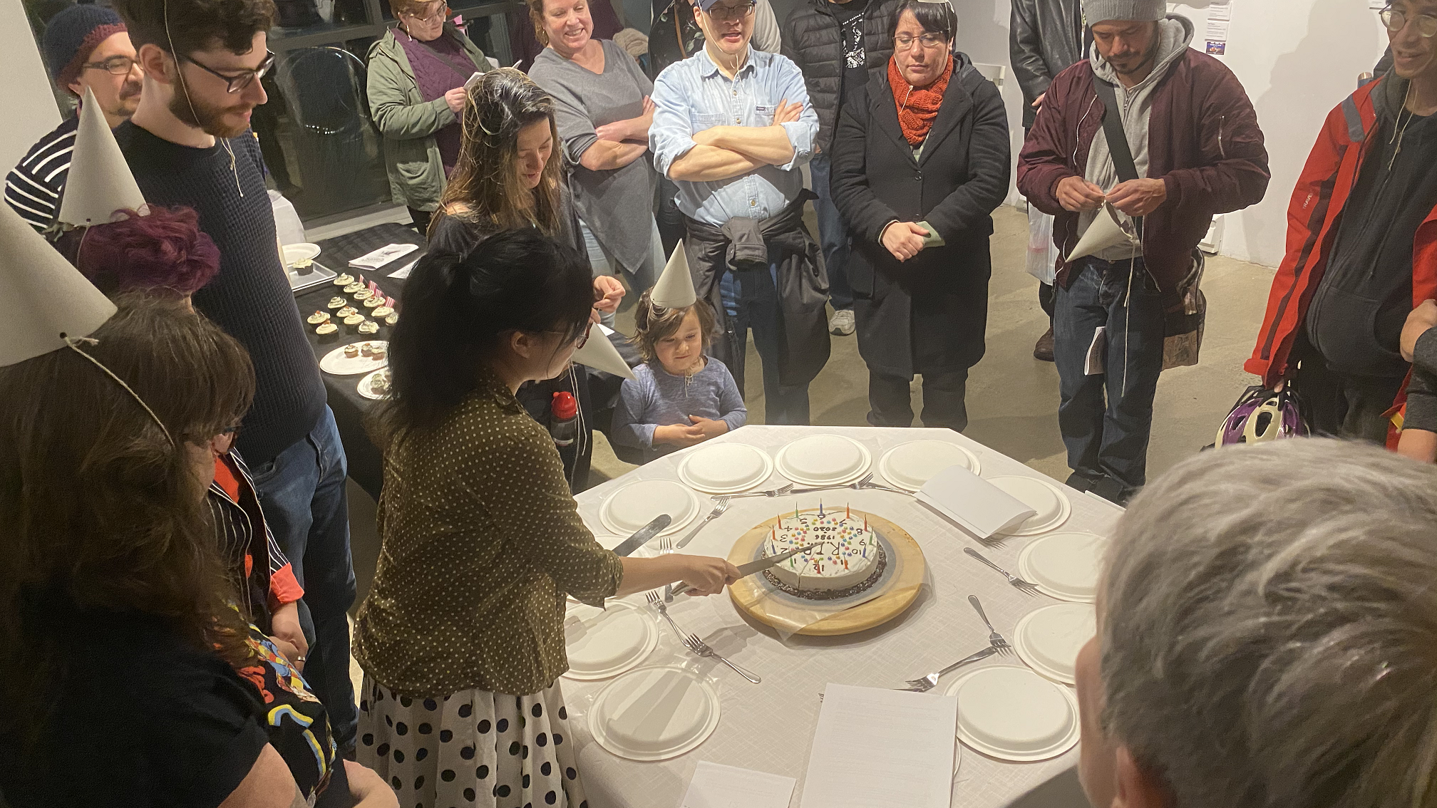 A large banner image of a public, in-person event. The photo shows an artist doing performance work with an audience around a round table.